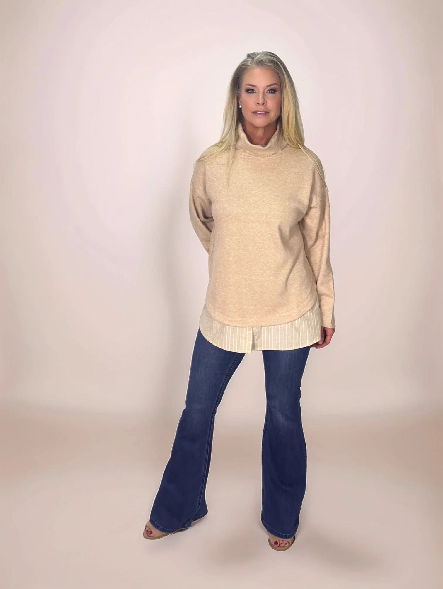 a blonde woman wearing a beige layered look turtleneck sweater with pinstriped shirt hem and dark blue jeans