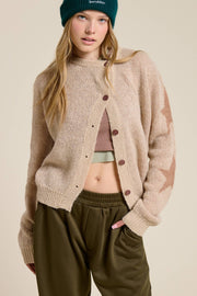 Woman wearing a beige cardigan, brown top, and green pants against a beige background
