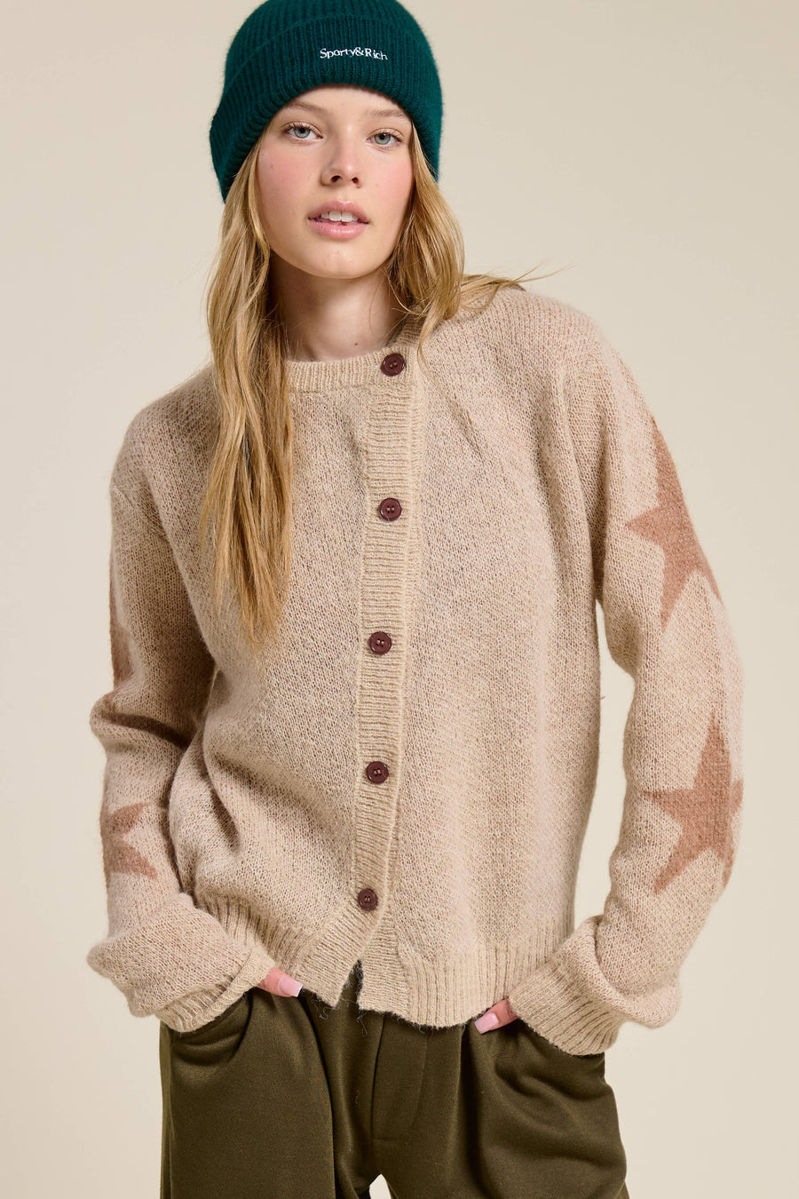 Woman wearing a beige cardigan with brown buttons and a teal beanie on a beige background