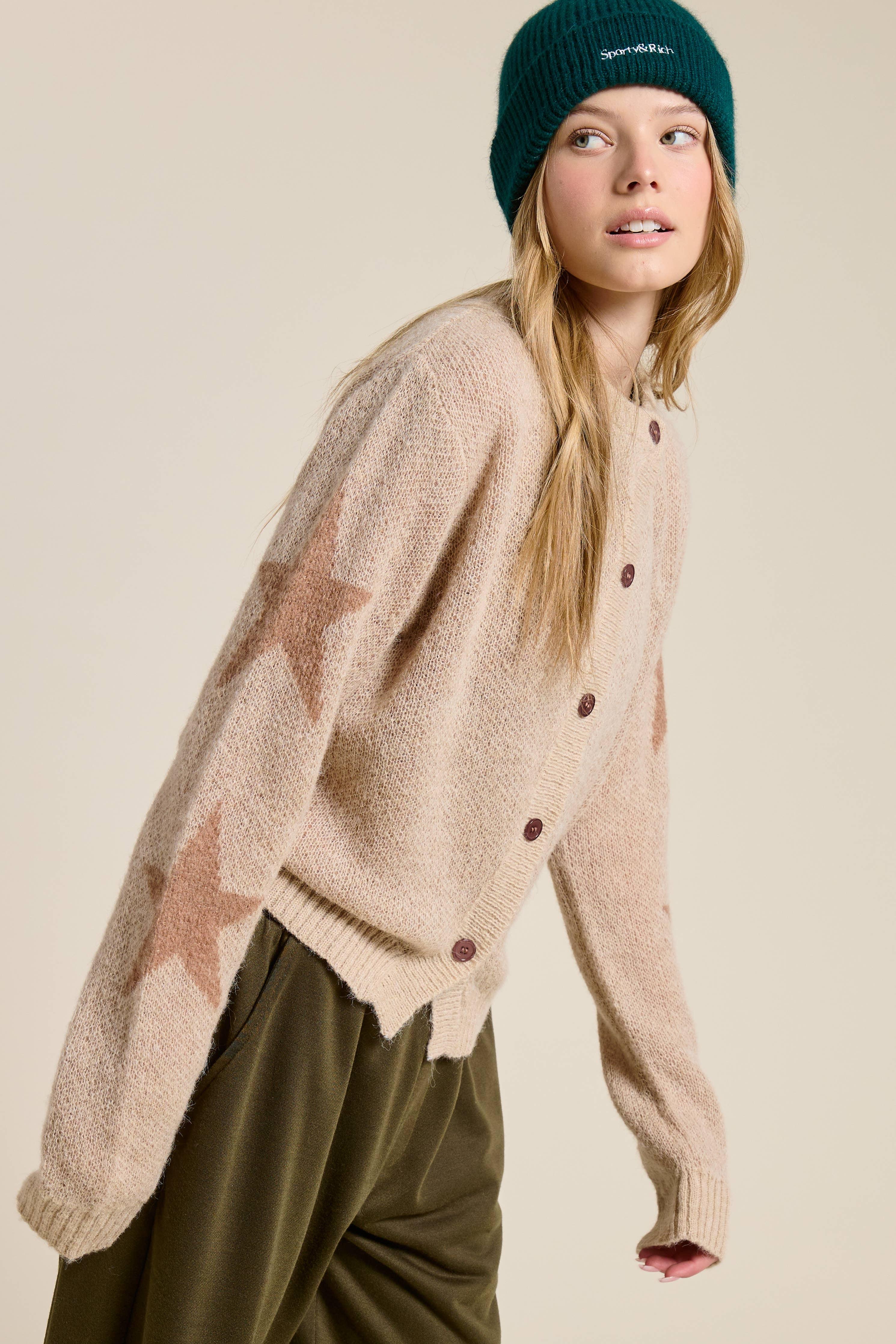 Woman wearing a beige sweater that has stars on the sleeves with brown buttons and a green beanie on a beige background
