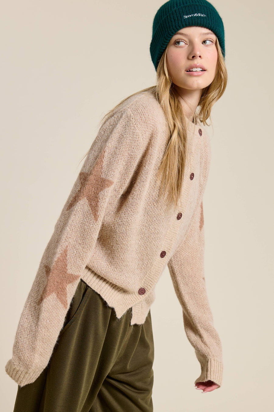 Woman wearing a beige sweater that has stars on the sleeves with brown buttons and a green beanie on a beige background