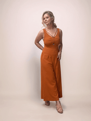 A blonde woman wearing a terracotta colored jumpsuit that  has a cutout at the bust and a v neckline. It features a ruched bust and added detailing strap at the neckline.