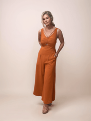 A blonde woman wearing a terracotta colored jumpsuit that  has a cutout at the bust and a v neckline. It features a ruched bust and added detailing strap at the neckline. The woman has one hand in her pocket and is wearing nude wedge sandals.