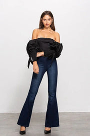 Woman wearing a black off-shoulder top and blue flared jeans is facing forward against a white background
