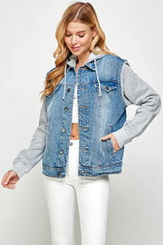 Blonde woman wearing a denim vest over a gray hoodie with white jeans and white crop top with one hand in her pocket.