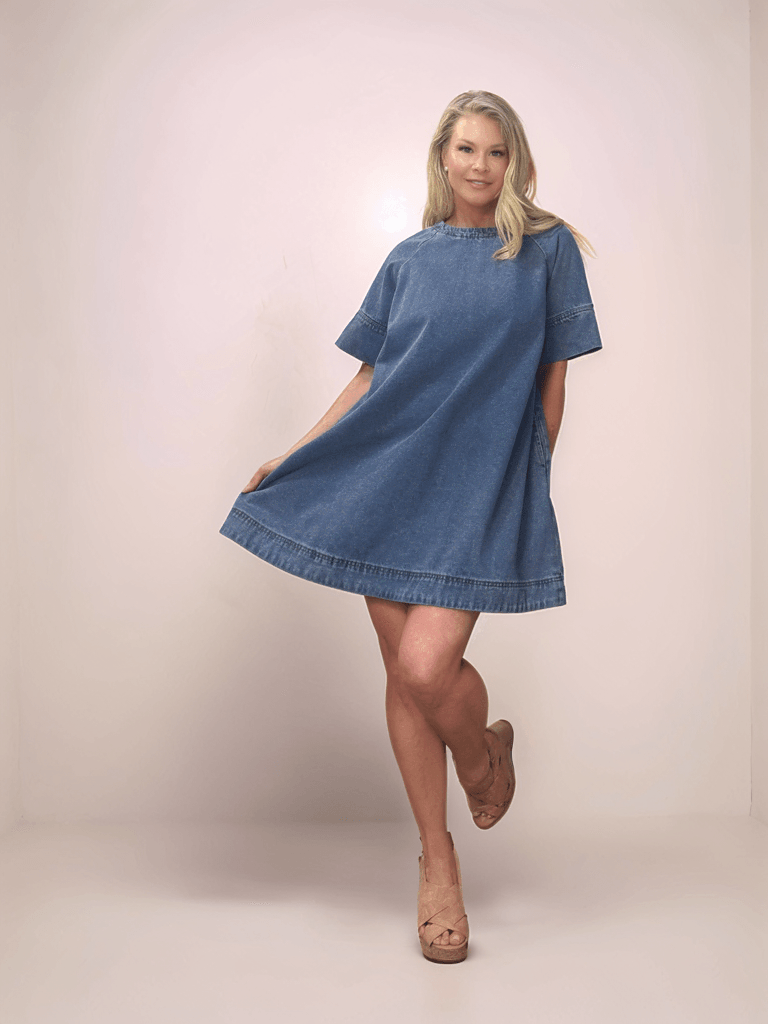 A blonde woman is smiling and wearing a medium blue a-line denim dress. She is holding the bottom of the dress to one side with one hand and has the other hand in her pocket.