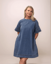 A blonde woman wearing a medium blue, loose fitting, a-line dress. She has both hands in her pockets. This is a perfect dress for casual events.