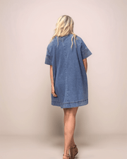 a blonde woman showing the back of a medium blue denim dress. The dress is loose fitting, with an a-line silhouette. It has elbow length sleeves and is above the knee.