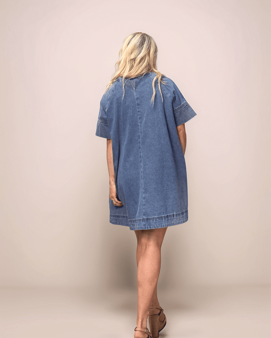 a blonde woman showing the back of a medium blue denim dress. The dress is loose fitting, with an a-line silhouette. It has elbow length sleeves and is above the knee.