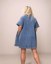 A blonde woman wearing a loose fitting, a line denim dress in medium blue. She has her hand in her pocket.