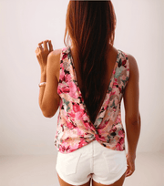 a woman with brown hair is wearing a floral sleeveless top that has a twist at the bottom of the open back. it is paired with white jean shorts.