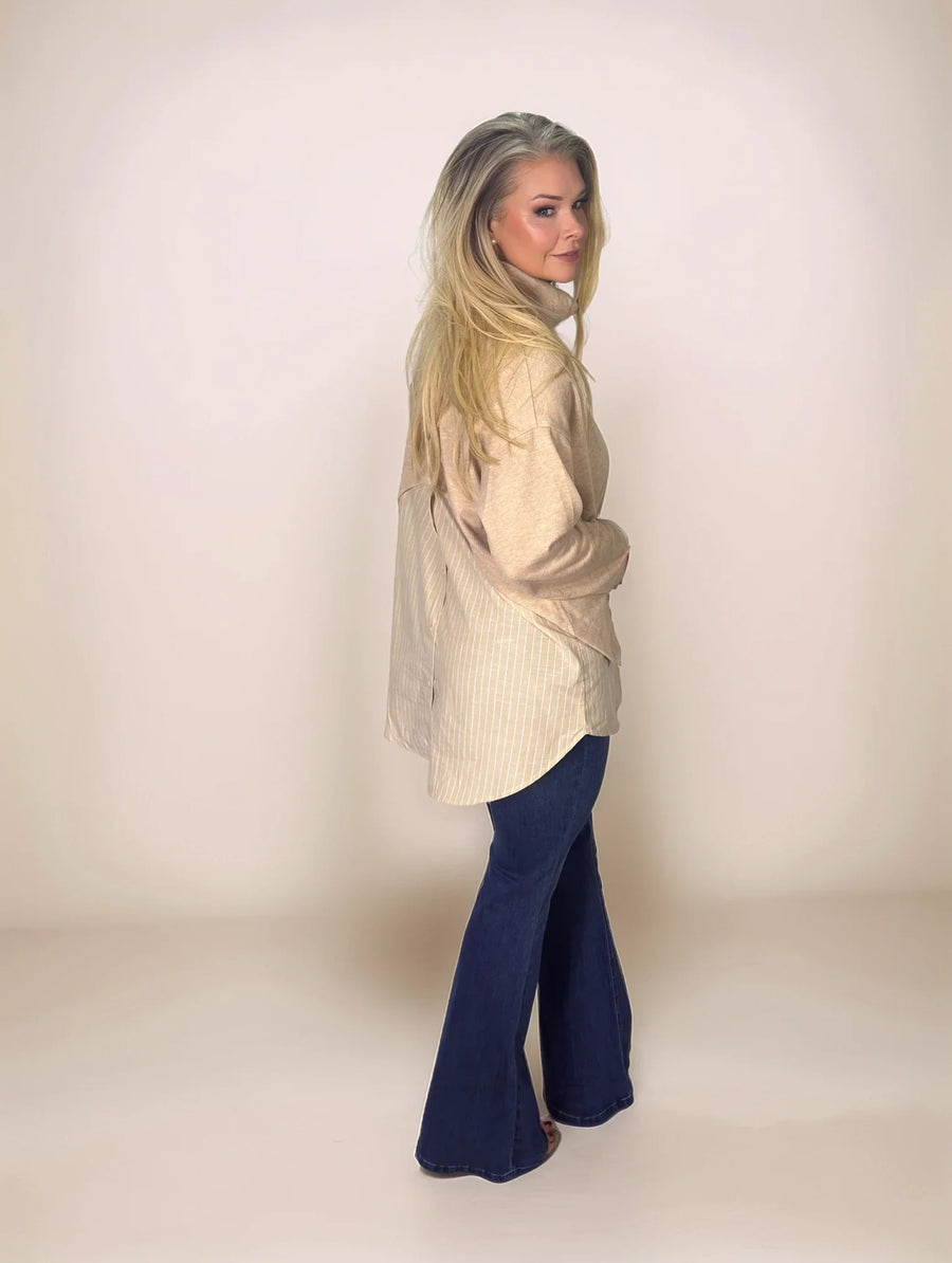 a blonde woman looking back towards the camera in a layered look beige sweater with a pinstripe long hem in the back and shorter hem in the front.