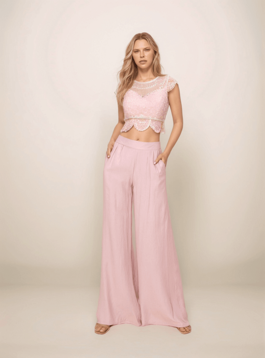 a blonde woman is standing with her hands in her pockets and is wearing pink wide leg pants and a cropped pink lace top