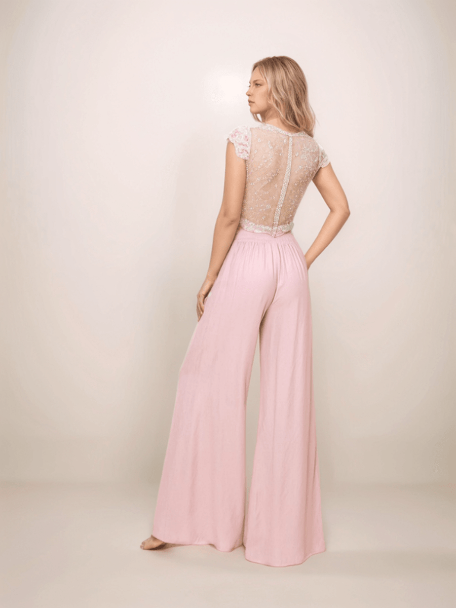 a blonde woman is standing facing backwards with one hand on her hip wearing pink wide leg pants and a sheer lace back top