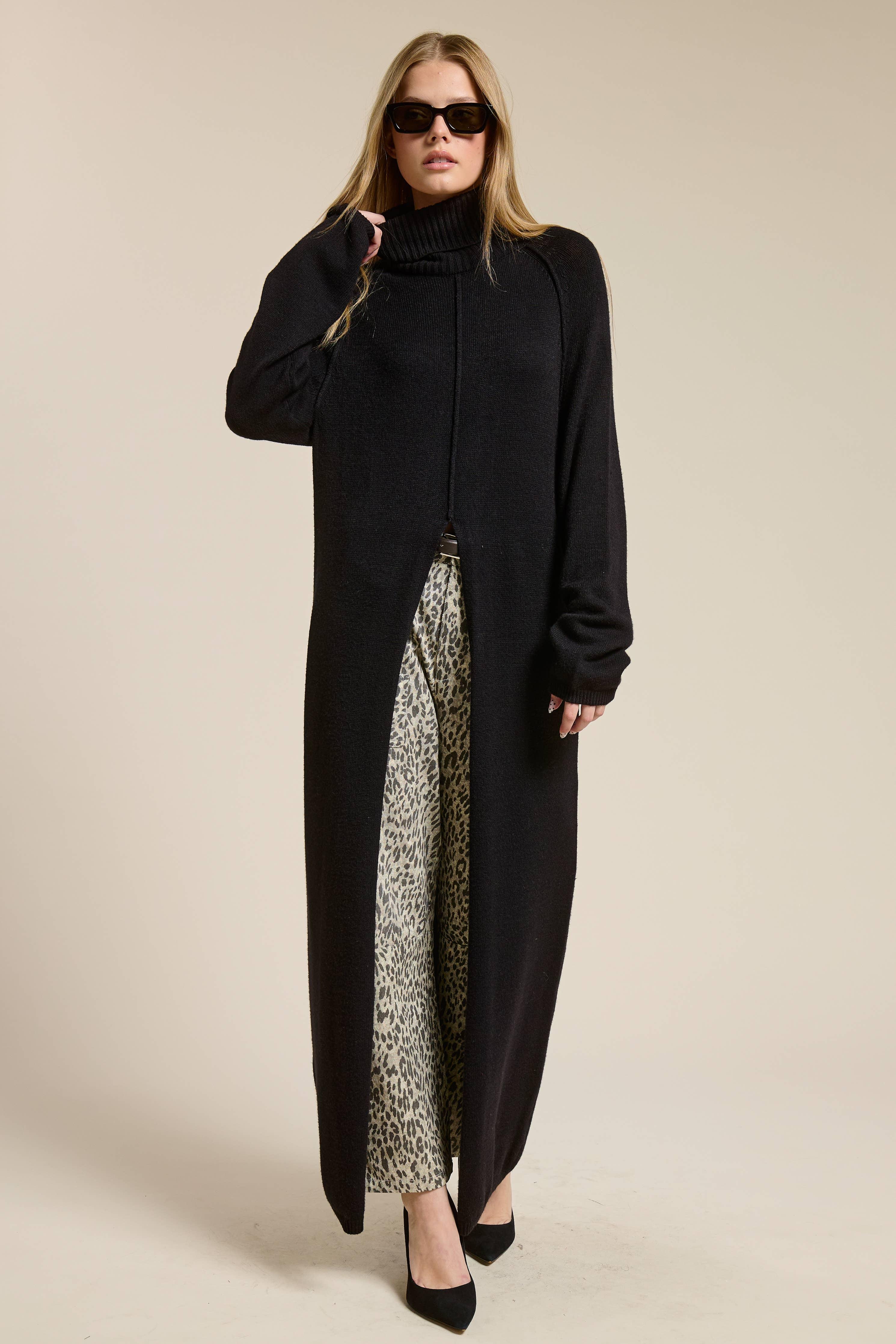 Woman wearing a long black sweater with a slit over patterned pants on a beige background