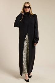 Woman wearing a long black sweater with a slit over patterned pants on a beige background