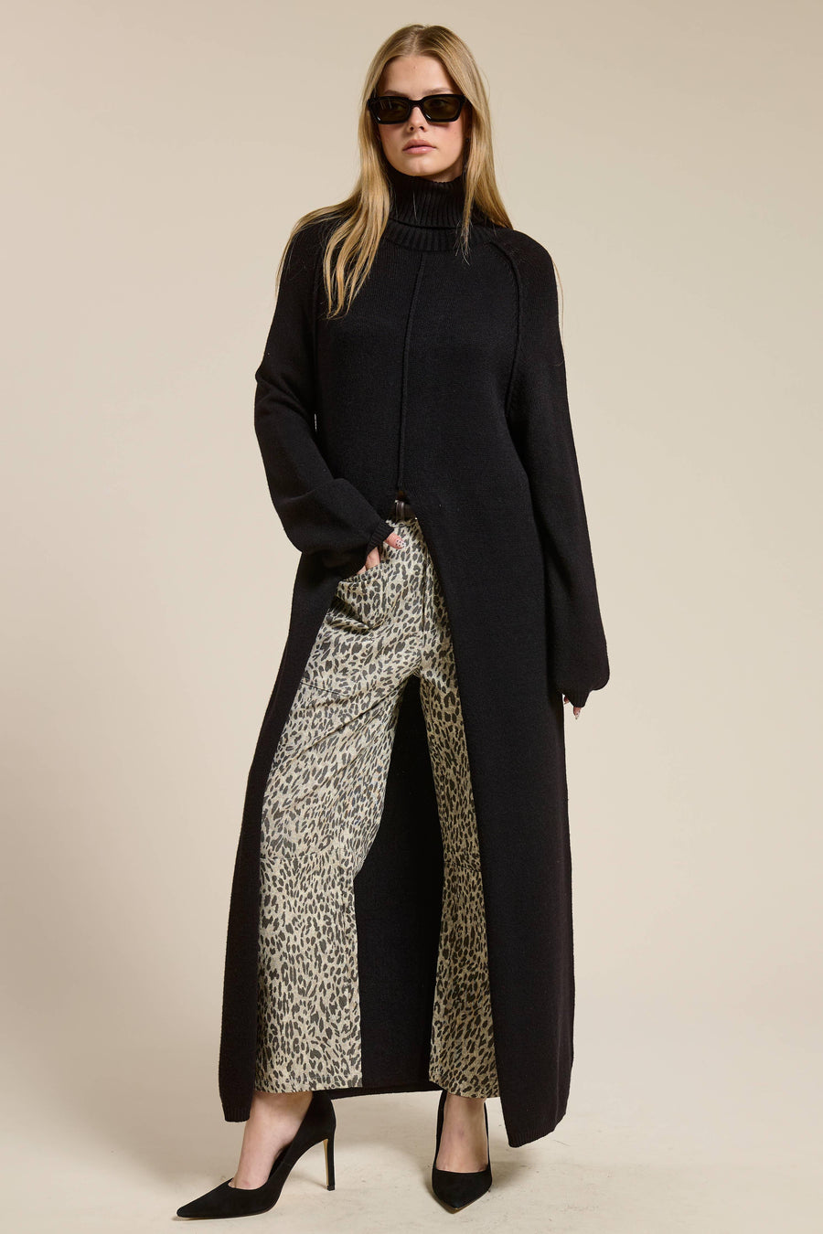Woman wearing a long black sweater with a slit in the middle over a patterned pair of pants on a beige background