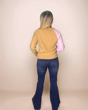 a blonde woman wearing dark denim jeans is facing away from the camera and is also wearing a camel colored sweater with one pink sleeve on the right.