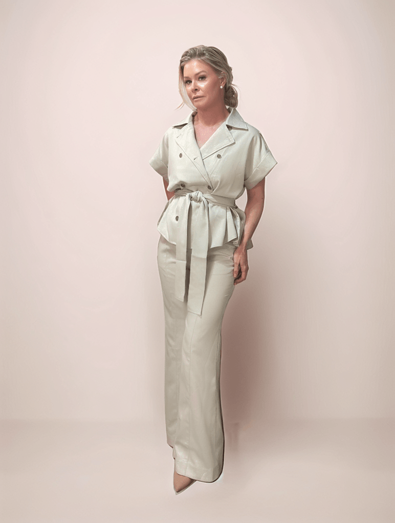 A blonde woman is wearing a matching two piece set in a beige color that features a pair of boot cut trousers and a belted double breasted jacket.