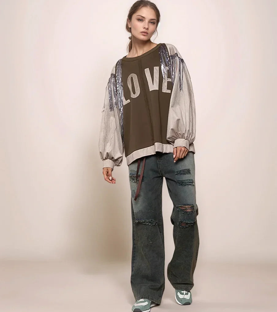 Woman wearing a brown 'LOVE' shirt with sheer sleeves and ripped jeans on a beige background