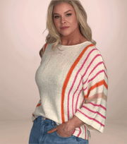 a blonde woman facing to the side and wearing a cream colored lightweight sweater that has orange and pink stripes from mid chest and down the arms. The sweater has a drop shoulder and wide sleeves. She has it paired with jeans