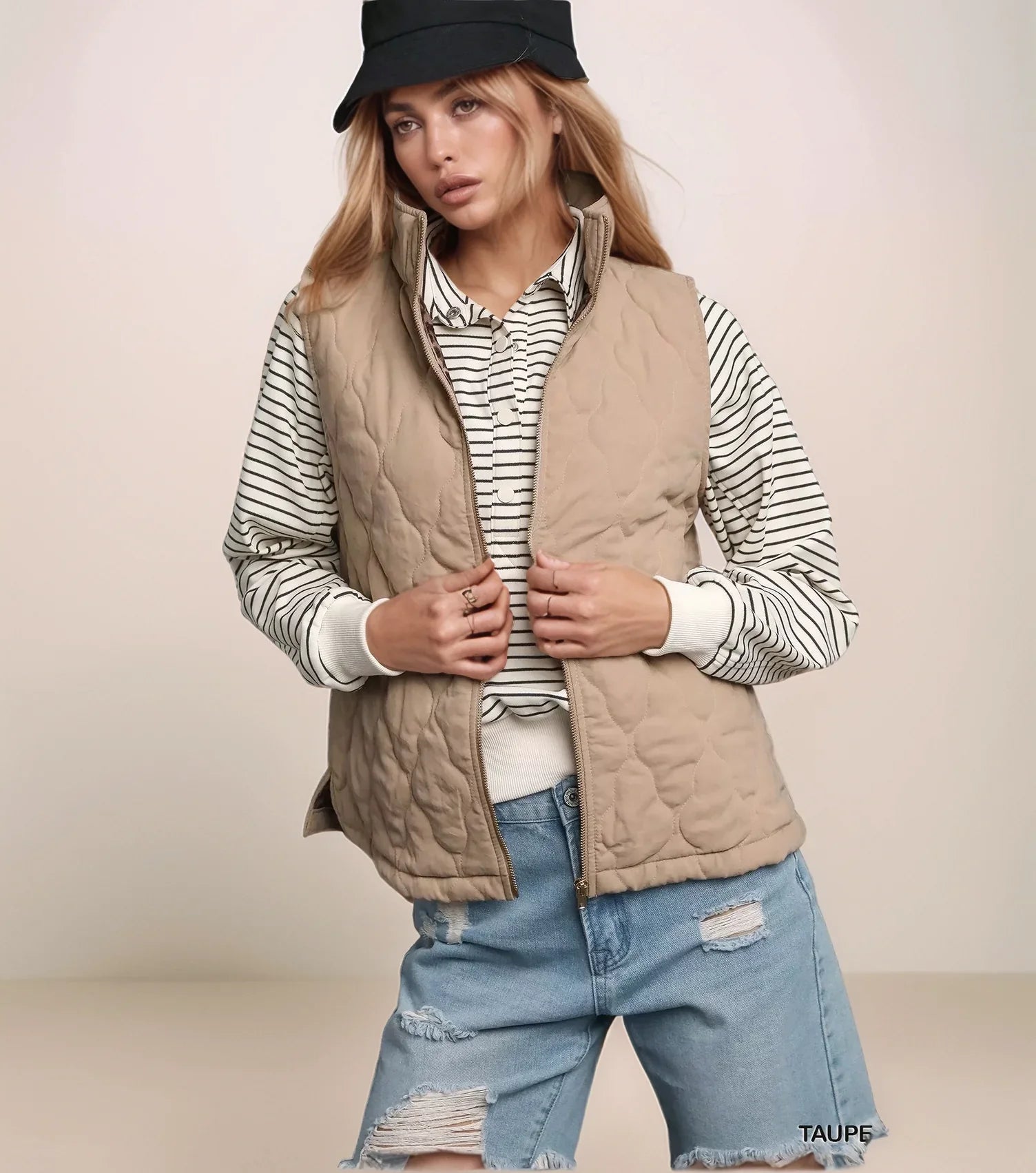 Woman wearing a beige quilted vest over a striped shirt with ripped jeans on a plain background