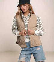Woman wearing a beige quilted vest over a striped shirt with ripped jeans on a plain background