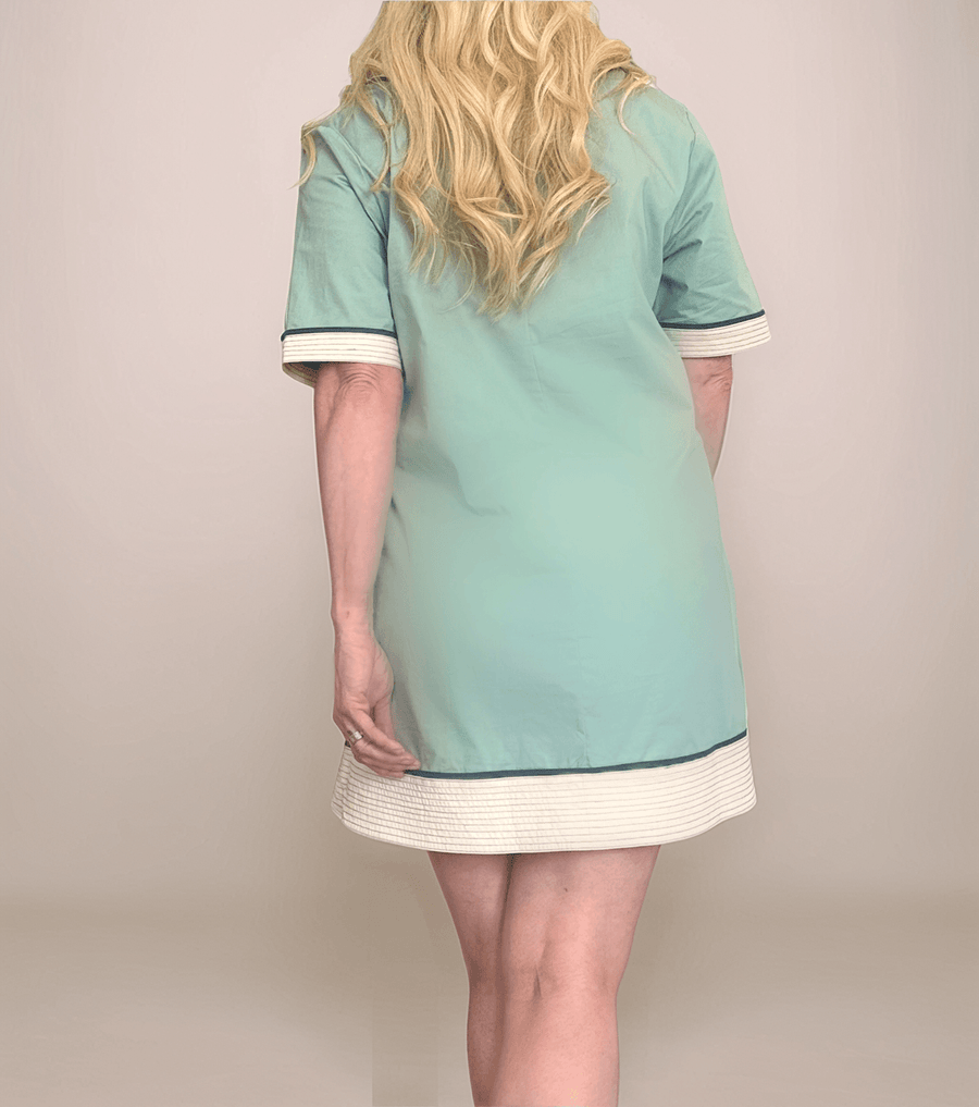 A confident blonde woman is wearing a retro-inspired, light slate colored tunic dress with cream and navy trim. She is wearing tan open-toe sandals.