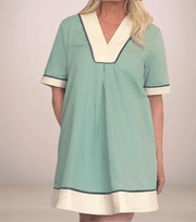 A confident blonde woman is wearing a retro-inspired, light slate colored tunic dress with cream and navy trim. She is wearing tan open-toe sandals.