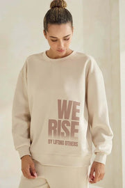 Person wearing a beige sweatshirt with 'WE RISE BY LIFTING OTHERS' text on a plain background
