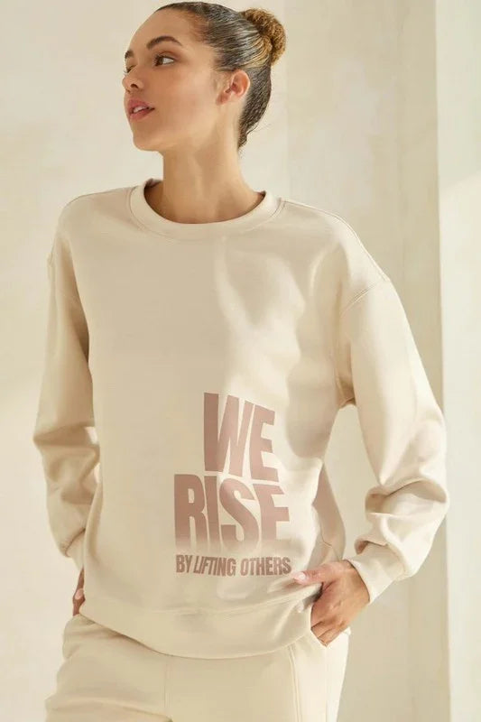 Woman wearing a beige sweatshirt with 'WE RISE BY LIFTING OTHERS' text on a plain background