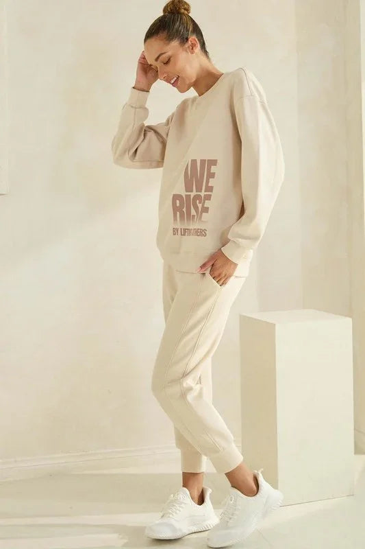 Woman wearing a beige 'WE RISE' sweatshirt and pants in a minimalistic setting