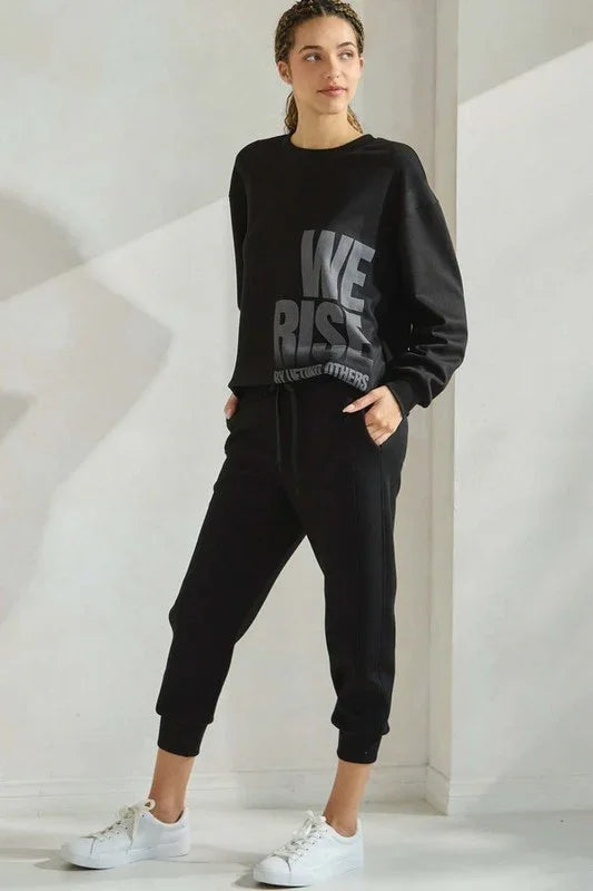 Person wearing a black 'WE RISE' sweatshirt and pants in a minimalistic setting