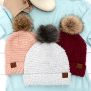 Three knitted hats with pom-poms in pink, white, and red on a light blue fabric background.