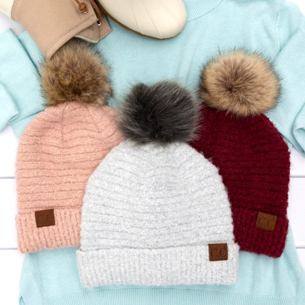 Three knitted hats with pom-poms in pink, white, and red on a light blue fabric background.