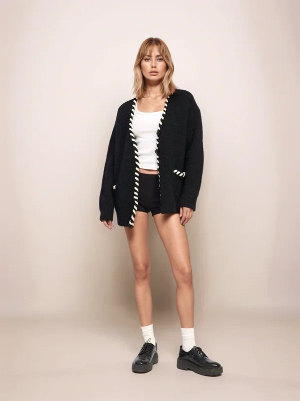 Woman wearing a black cardigan with white accents, white top, black shorts, and black shoes on a beige background