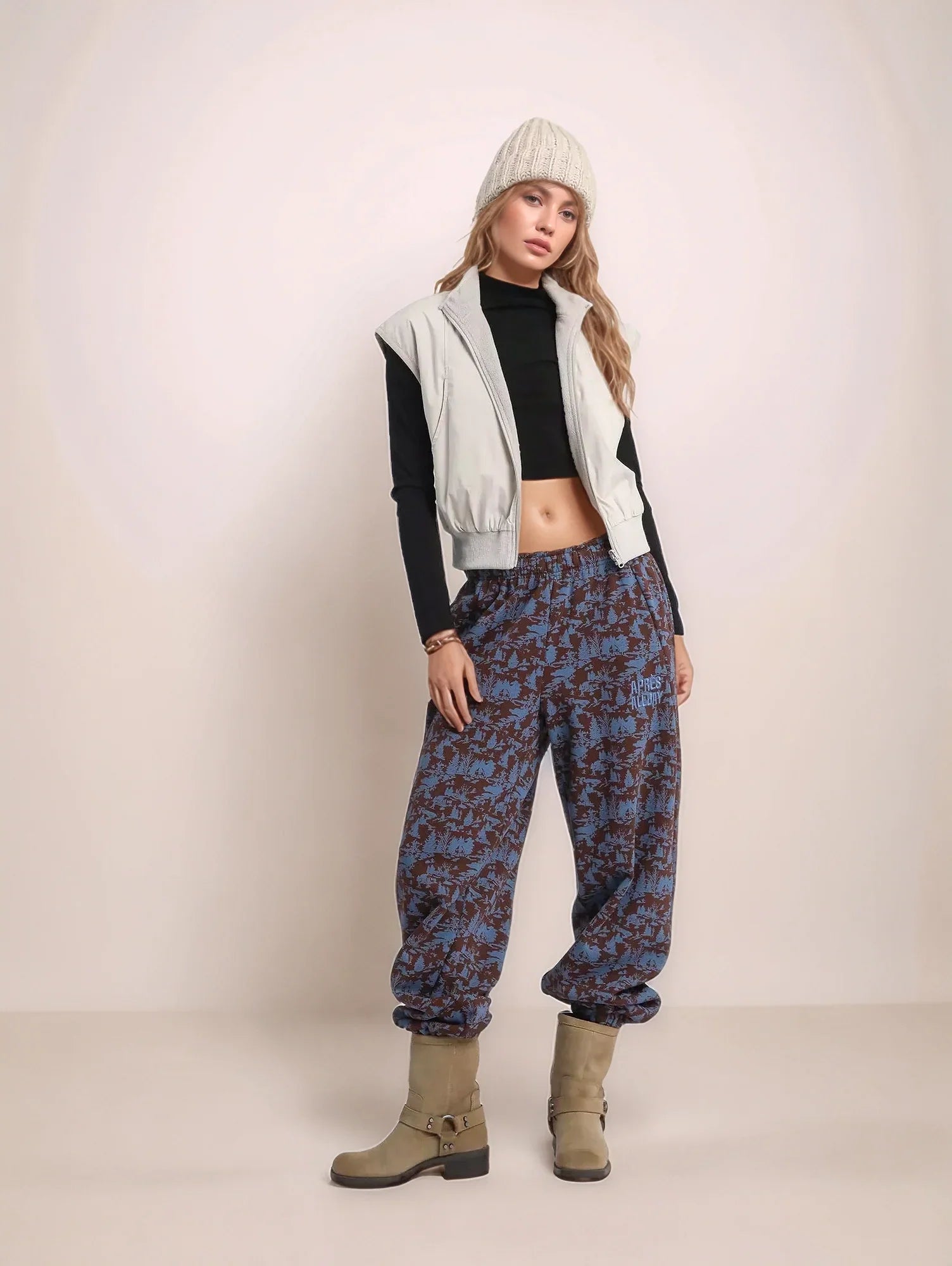 Person wearing a black crop top, white vest, blue patterned pants, and beige boots on a plain background