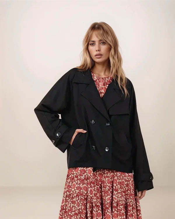 Woman wearing a black cropped trench coat over a floral dress on a plain background