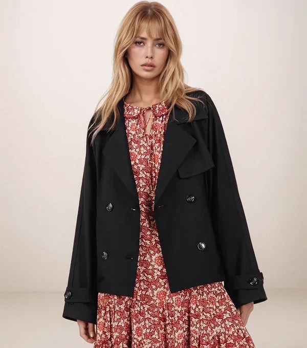 Woman wearing an open black cropped trencb coat over a red floral dress on a plain background