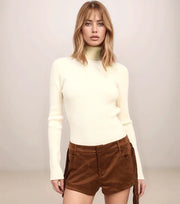 Woman wearing a white turtleneck and brown shorts against a plain background