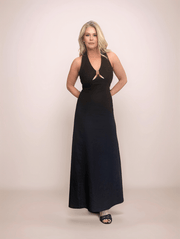 a blonde woman standing with her hands behind her back wearing a black linen maxi dress. the dress has a halter neck and a v neck with a cutout at the bust held together by gold hardware. The bodice is fitted and the bottom of the dress is a line style.