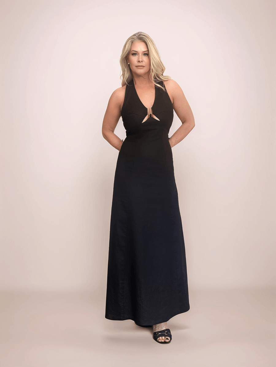 a blonde woman standing with her hands behind her back wearing a black linen maxi dress. the dress has a halter neck and a v neck with a cutout at the bust held together by gold hardware. The bodice is fitted and the bottom of the dress is a line style.