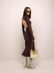 Woman wearing a brown dress with flared sleeves and patterned pants, holding a straw bag against a white background.