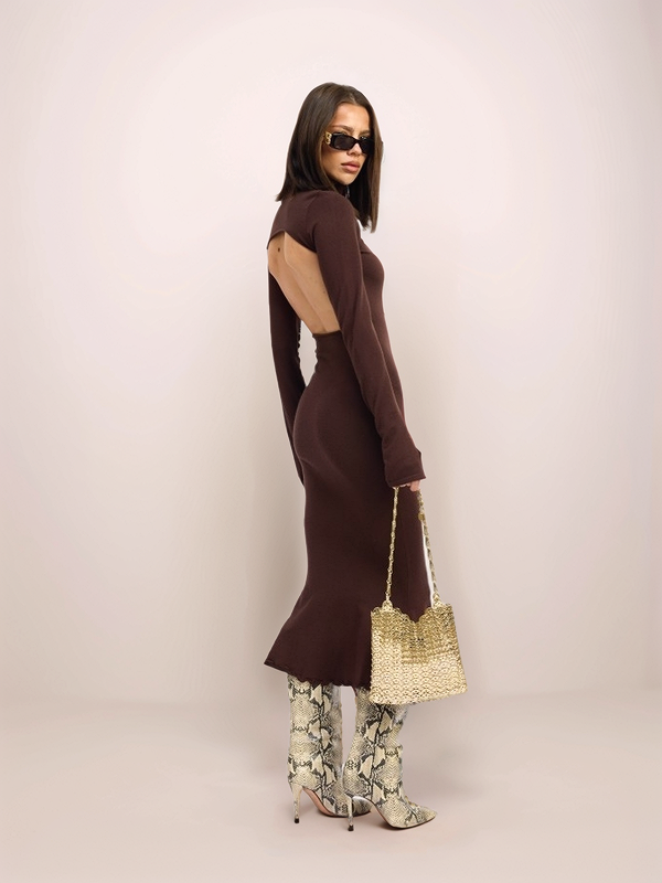 Woman wearing a brown dress with flared sleeves and patterned pants, holding a straw bag against a white background.