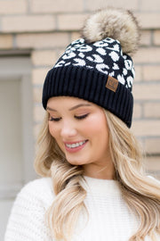 Woman wearing a black and white patterned beanie with a fur pom-pom against a brick wall.