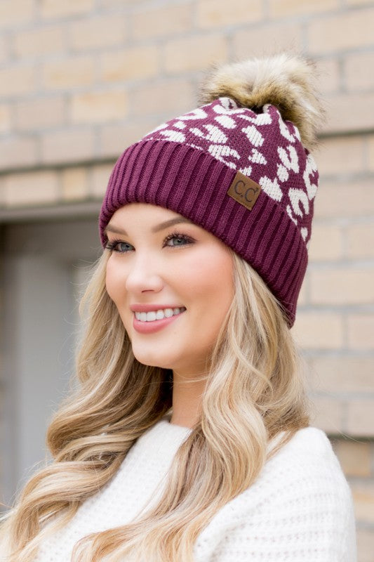 Woman wearing a maroon beanie with a pom-pom against a brick wall.