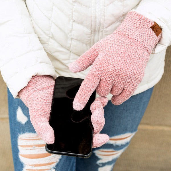 Person wearing pink fleece gloves holding a smartphone, with a white jacket and blue jeans in the background.