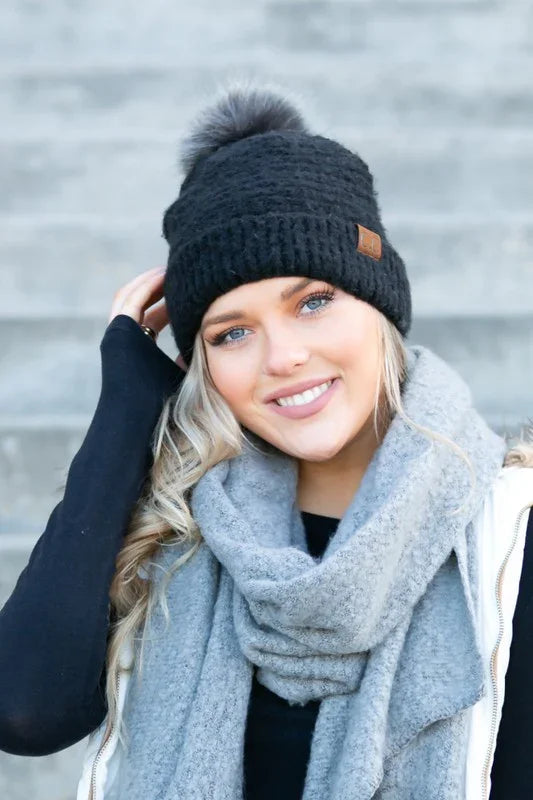 Woman wearing a black knit beanie with a pom-pom and a gray scarf against a blurred background