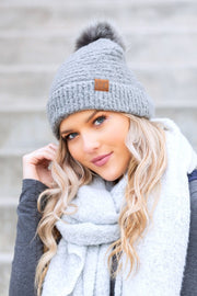 Woman wearing a gray knit beanie with a pom-pom and a white scarf against a neutral background
