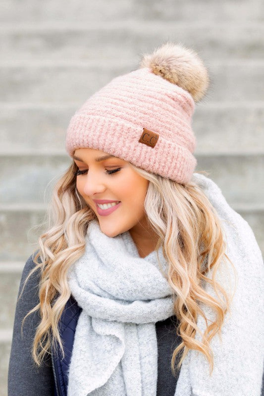 Woman wearing a pink knit beanie with a pom-pom and a gray scarf, standing against a neutral background.